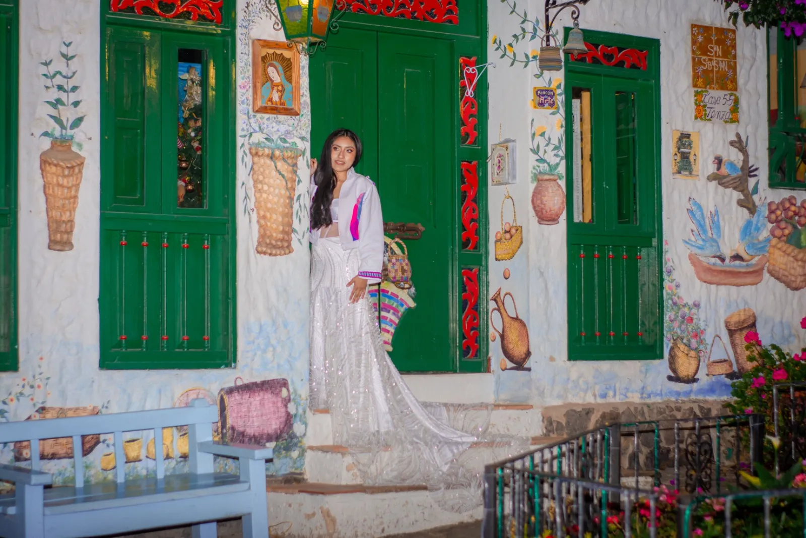Mujer con chaqueta blanca y vestido bordado junto a una fachada pintada