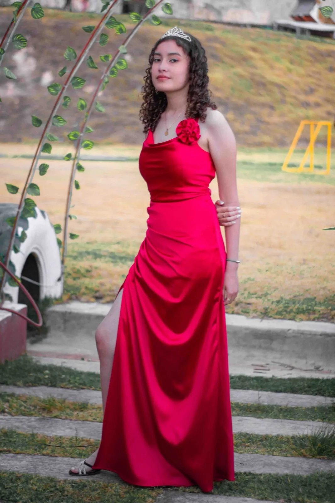 Quinceañera con vestido rojo de satín y corona sobre unas escaleras de jardín