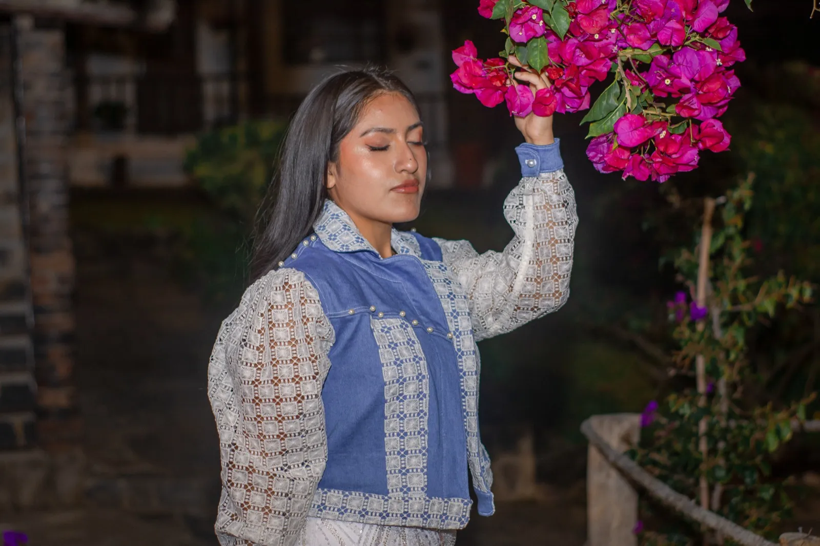 Mujer con chaleco de denim azul y chaqueta de encaje blanco levantando la mano hacia las buganvilias