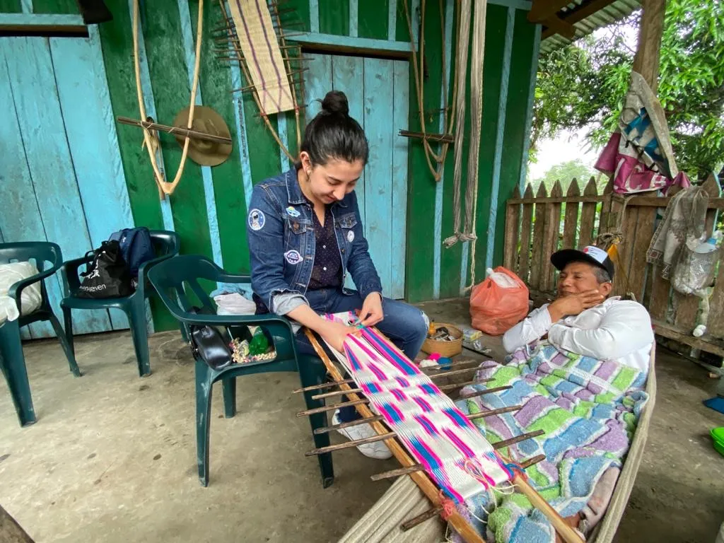 Mujer con chaqueta de denim tejiendo en un telar junto a un hombre recostado en una hamaca